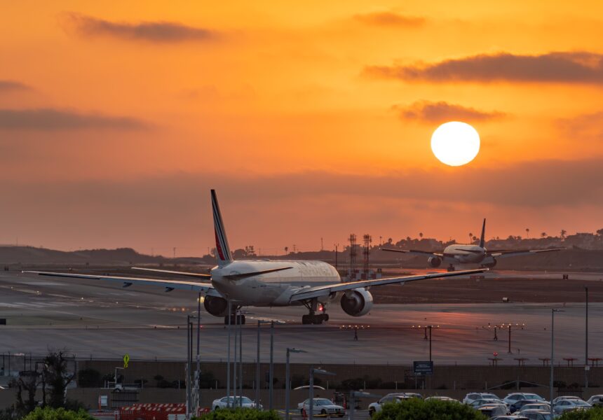 Two wide-body commercial passenger jetliners sit on an airport tarmac during a vivid, glowing orange sunset. The foreground aircraft is viewed from behind as it taxis down the runway, while another plane taxies further in the distance under a bright, low-hanging sun.