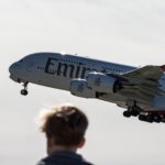 Low-angle view of an Emirates Airbus A380 passenger jet ascending into the sky with landing gear extended, seen from behind the blurred silhouette of a person watching the departure.