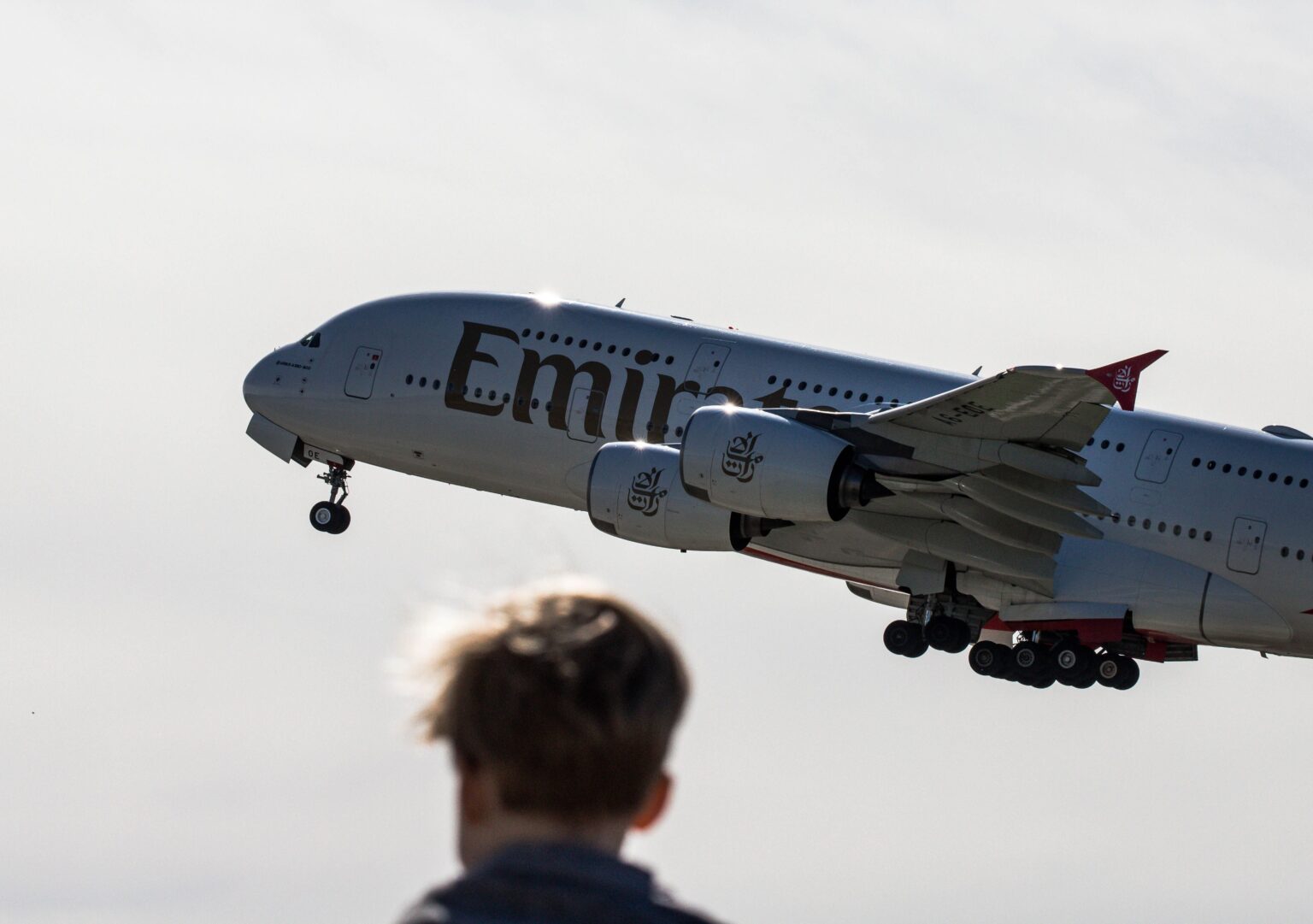 Low-angle view of an Emirates Airbus A380 passenger jet ascending into the sky with landing gear extended, seen from behind the blurred silhouette of a person watching the departure.