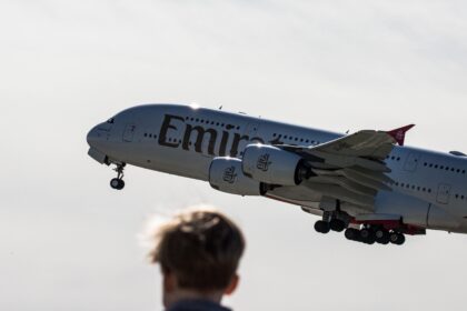 Low-angle view of an Emirates Airbus A380 passenger jet ascending into the sky with landing gear extended, seen from behind the blurred silhouette of a person watching the departure.
