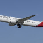 The photo shows a Qantas aircraft mid-flight, with a blue sky as the background.