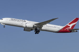 The photo shows a Qantas aircraft mid-flight, with a blue sky as the background.