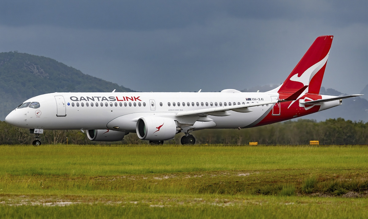 A QantasLink aircraft having just touched down on the runway. The runway is surrounded by grass. A mountain can be seen in the distance, behind the aircraft, as well as a grey sky.