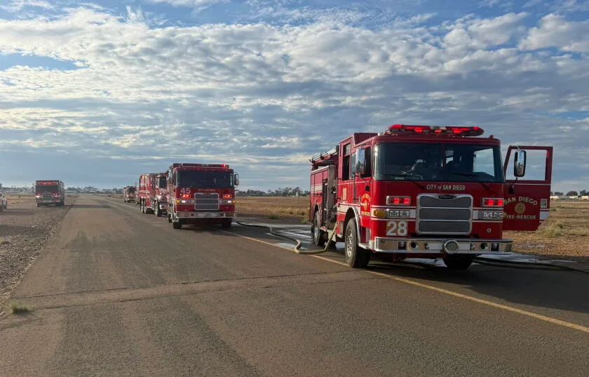 A brownish grey tarmac road occupies the bottom half of the image, with 5 red fir trucks lined up on it, getting further away from the camera further down he line. The San Diego Fire-Rescue Department logo can be seen on the side doors of the first vehicle in the line. The background of the image is pale blue sky with fluffy white clouds. 