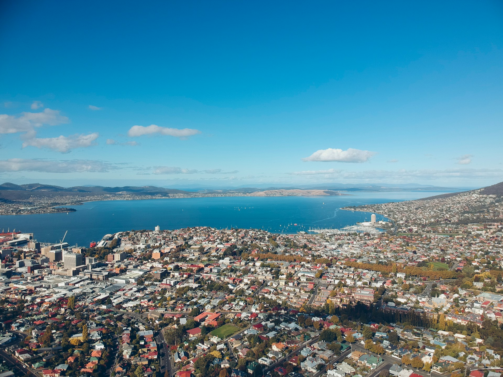 A view of Hobart from above, showing a view of buildings, houses and a body of water under blue skies with some clouds
