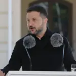 An image of Ukrainian President Volodymyr Zelensky giving an address with two microphones positioned in front of his face.