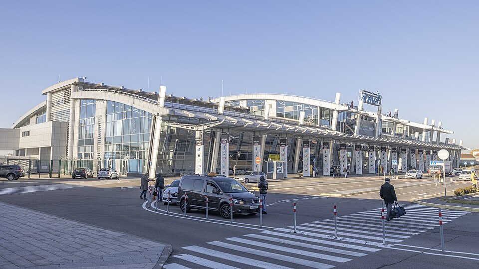 The exterior of Kiev's Zhuliany International Airport. The outside of the terminal building can be seen surrounded by a road being used by cars and pedestrians. 