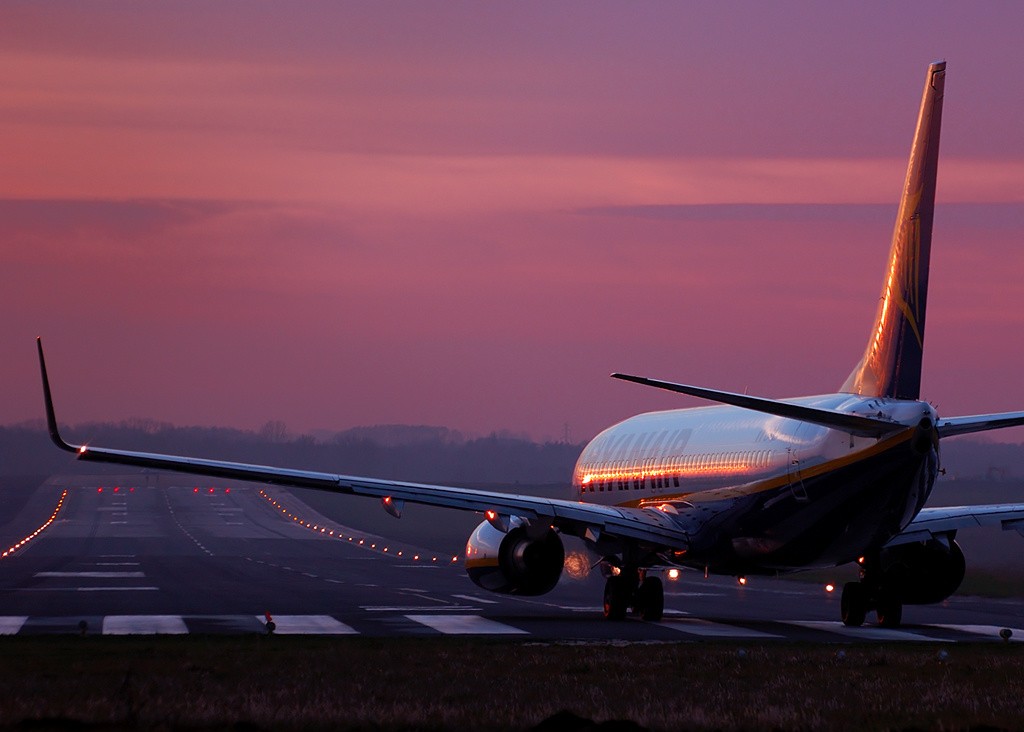 Ryanair Boeing 737 aircraft taxiing on a runway at dusk, with runway lights illuminated and a pink-purple sunset sky in the background.