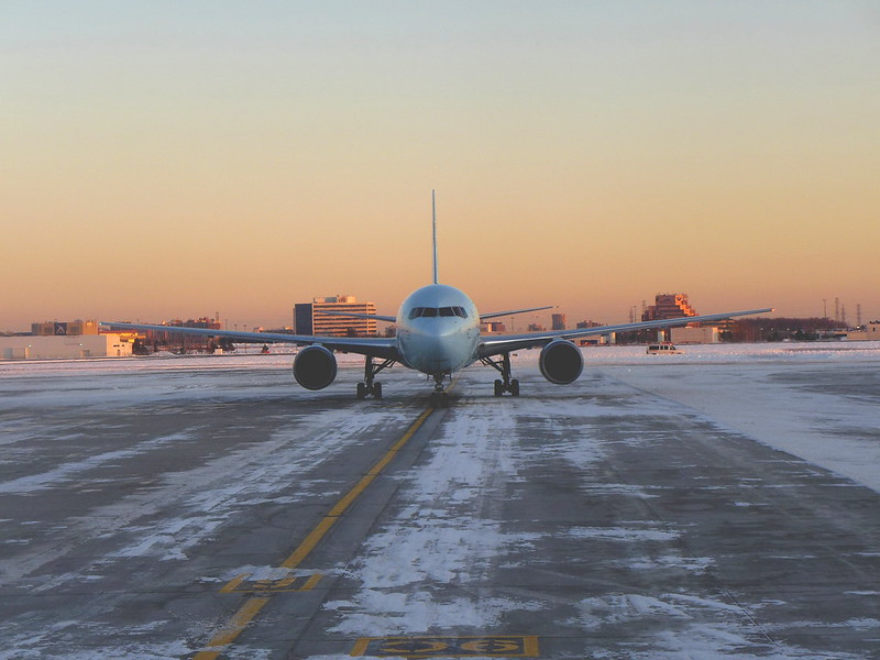 Twin-engine airliner seen head-on, taxiing along a snow-dusted yellow-lined taxiway under an orange and pale blue dusk sky in Toronto Airport.