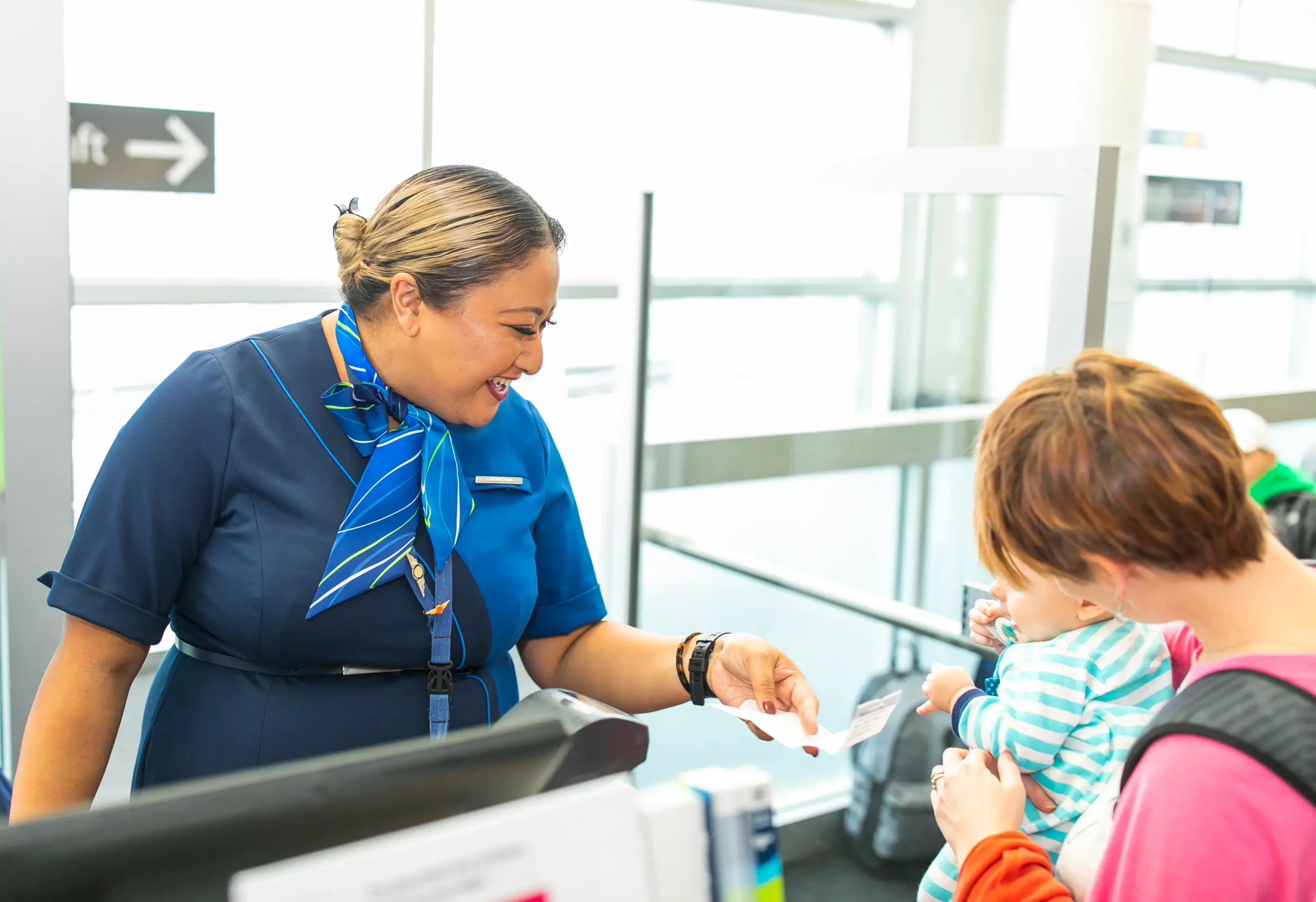 Smiling gate agent in a blue uniform and patterned scarf hands a boarding pass to a woman holding a baby in a striped top at an airport gate desk.