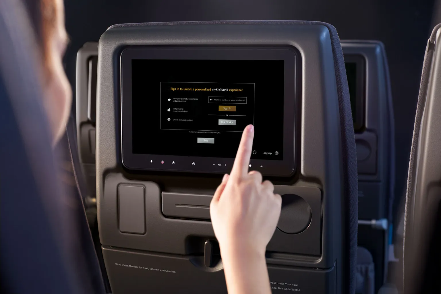 Passenger’s finger pointing at a seatback screen showing a sign-in page with Sign In, Pair Device and Skip buttons, above a cup holder.
