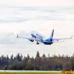 Photo depicting an Alaska Airlines aircraft taking off from an airport runway, heading north. It is overhead a wooded area, with a white, clouded sky in the background.