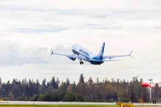 Photo depicting an Alaska Airlines aircraft taking off from an airport runway, heading north. It is overhead a wooded area, with a white, clouded sky in the background.