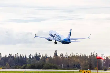 Photo depicting an Alaska Airlines aircraft taking off from an airport runway, heading north. It is overhead a wooded area, with a white, clouded sky in the background.