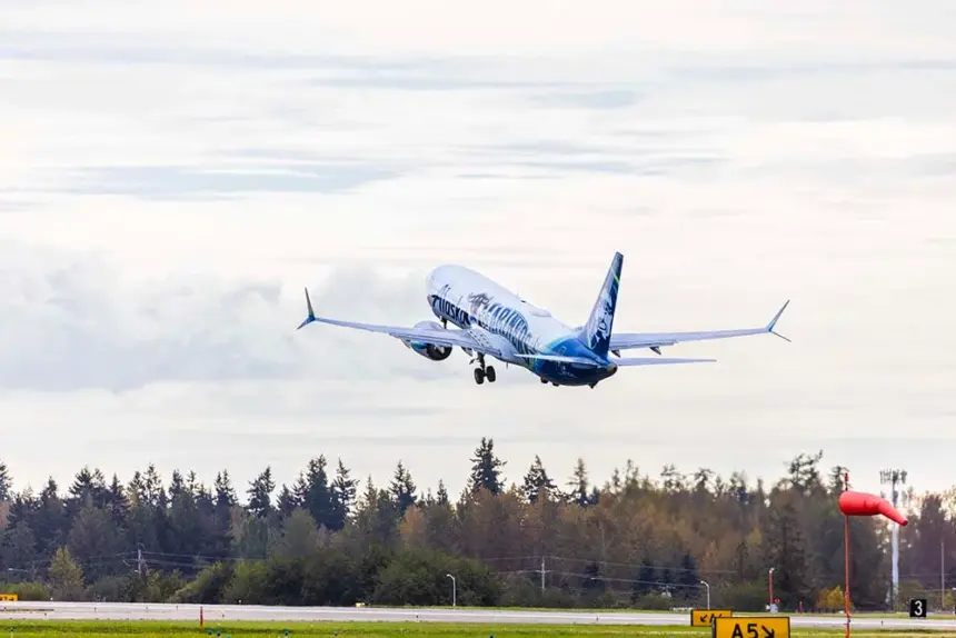 Photo depicting an Alaska Airlines aircraft taking off from an airport runway, heading north. It is overhead a wooded area, with a white, clouded sky in the background.