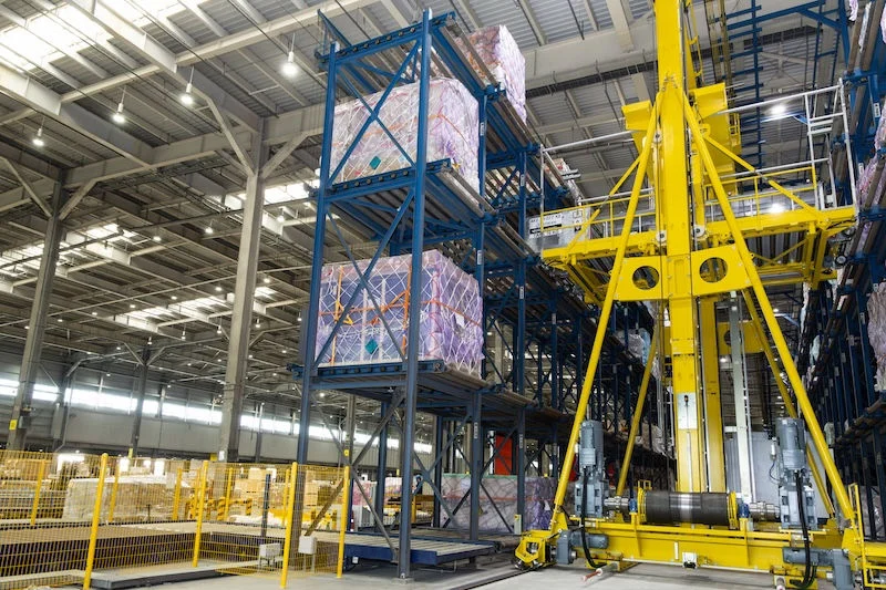 Inside a large warehouse, a tall yellow crane runs beside blue multi-level racking holding wrapped cargo pallets, with yellow safety fencing at left