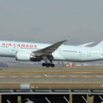 Air Canada Boeing 787-8 Dreamliner taking off, nose wheel raised, with a red maple leaf on a pale turquoise tail, seen above a fence and runway.