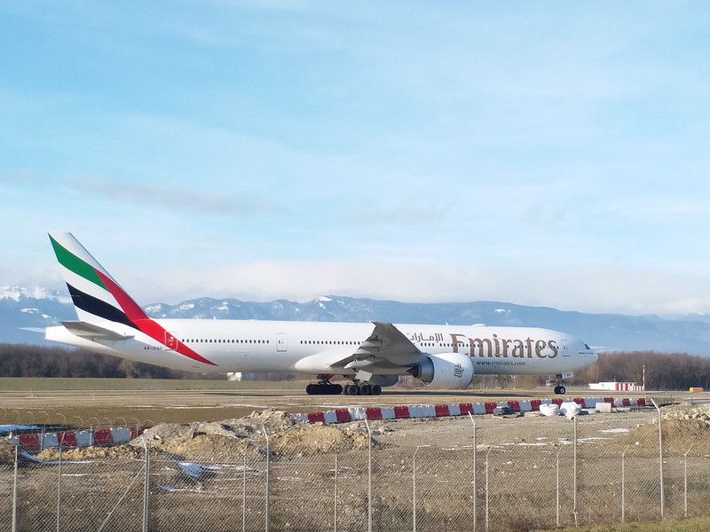 White wide-bodied jet on a runway beyond a wire fence and red-and-white barriers, with a red, green and black striped tail and snowy hills under a pale sky.
