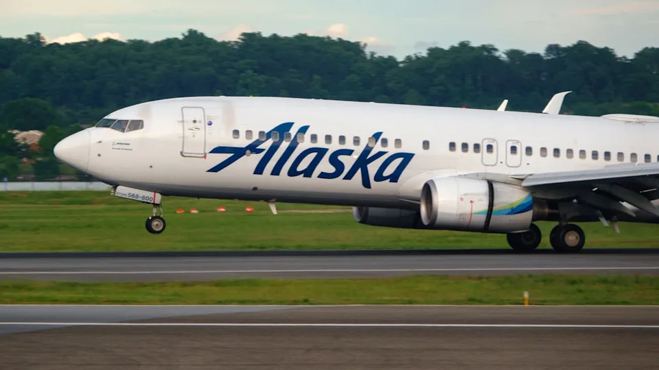 Photo of an Alaska Airlines aircraft initiating takeoff from an airport runway. Trees and a blue sky can be seen in the background.