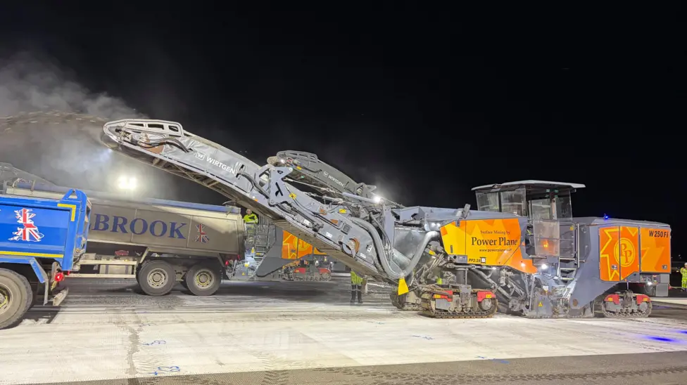 Image shows a large milling machine and other vehicles on the runway at Heathrow Airport at night