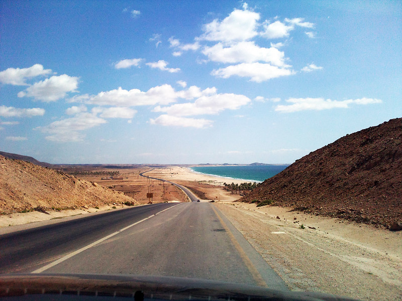 View through a car windscreen of a road descending between barren brown hills towards a turquoise sea, with palm trees and a sandy coast ahead.