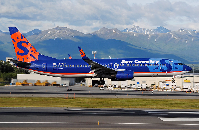 Blue jet with red and orange trim and a large orange sunburst on the tail, wheels just touching a runway, with snow-capped mountains and cloud behind.