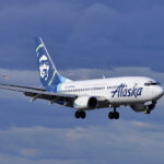 Alaska Air jet on approach with landing gear extended under a blue, partially cloudy sky, with a blue tail. A smiling Inupiat profile is featured on the aircraft's tail.