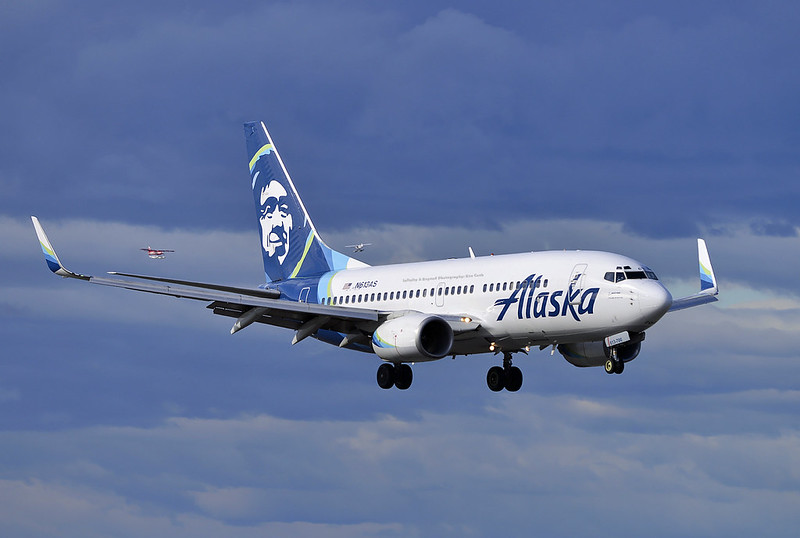 Alaska Air jet on approach with landing gear extended under a blue, partially cloudy sky, with a blue tail. A smiling Inupiat profile is featured on the aircraft's tail.