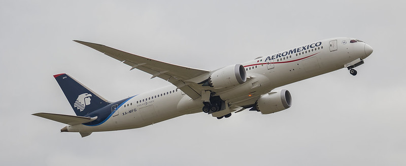 Twin-engine jet climbing against an overcast sky, landing gear still down, with a blue tail bearing a white eagle emblem and a red stripe.