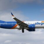 Jet on approach with landing gear down against a cloudy blue sky, with a white front fuselage, blue rear section and an orange sunburst design on the tail.