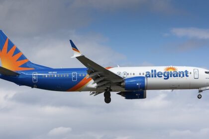 Jet on approach with landing gear down against a cloudy blue sky, with a white front fuselage, blue rear section and an orange sunburst design on the tail.