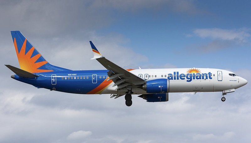 Jet on approach with landing gear down against a cloudy blue sky, with a white front fuselage, blue rear section and an orange sunburst design on the tail.