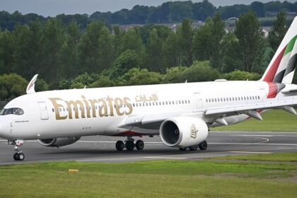 White wide-bodied jet taxiing on a runway with gold lettering in English and Arabic, a tail striped in red, green and black, and trees behind.