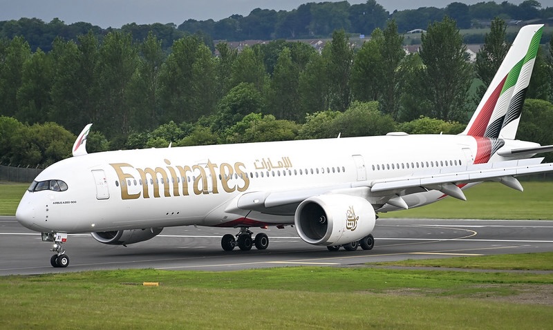 White wide-bodied jet taxiing on a runway with gold lettering in English and Arabic, a tail striped in red, green and black, and trees behind.