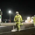 Image shows a team of maintenance workers in yellow suits and safety equipment spraying the tarmac at well-lit Heathrow Airport at night with a truck in the background