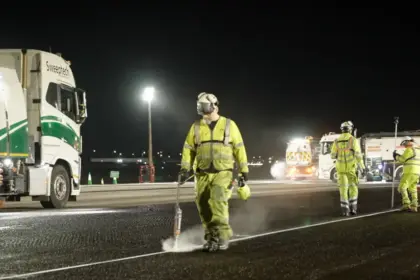 Image shows a team of maintenance workers in yellow suits and safety equipment spraying the tarmac at well-lit Heathrow Airport at night with a truck in the background