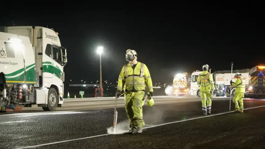 Image shows a team of maintenance workers in yellow suits and safety equipment spraying the tarmac at well-lit Heathrow Airport at night with a truck in the background