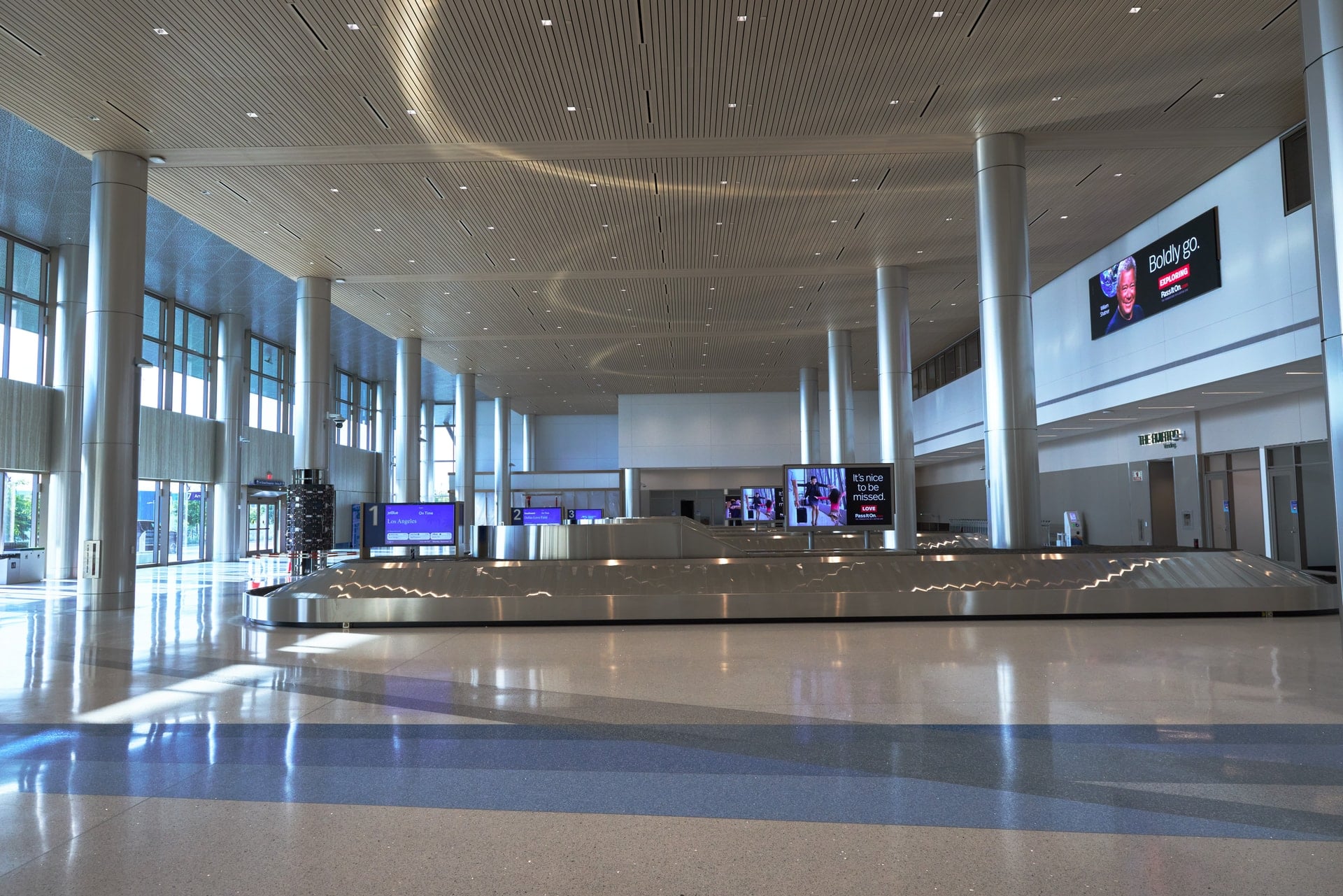 A spacious, modern airport baggage claim area with polished floors, tall metal columns, a slatted ceiling, and digital displays above the conveyors.