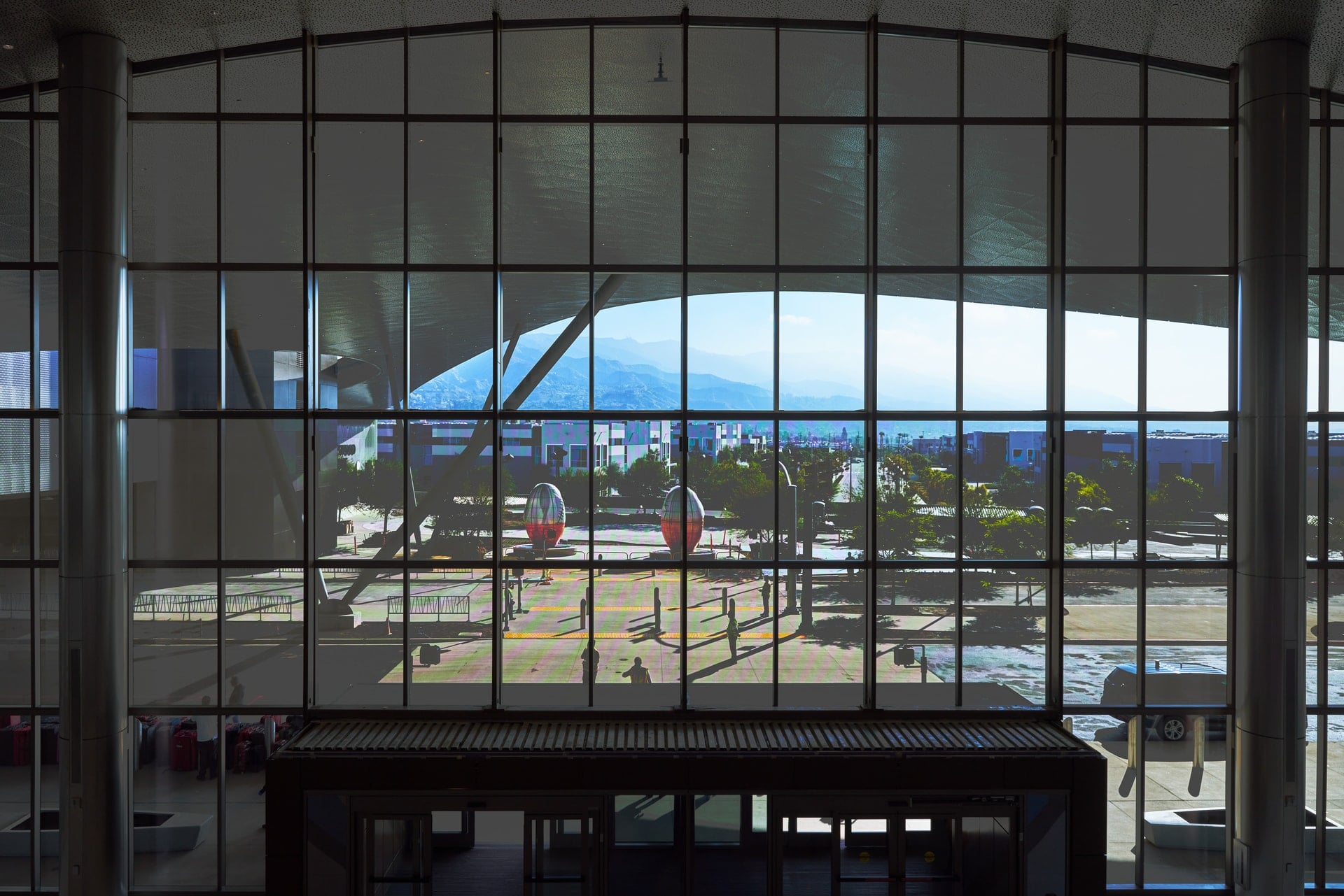 A view through a large glass wall of the new terminal in Hollywood Burbank Airport, showing an outdoor plaza with two red sculptures, trees, and distant mountains under a sky.