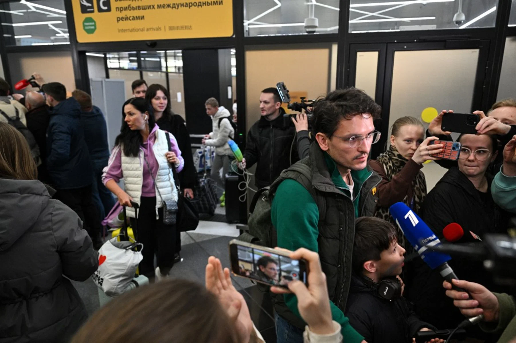 Passengers arrive at Sheremetyevo International Airport with journalist microphones and mobile phones surrounding them.
