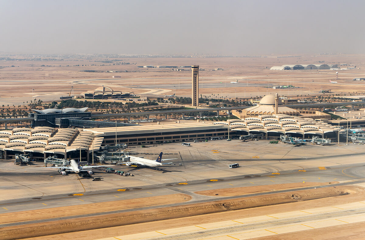 Aerial view of King Khalid International Airport (RUH) and control tower surrounded by runways and taxiways, with several commercial aircraft parked at the gates in a desert landscape