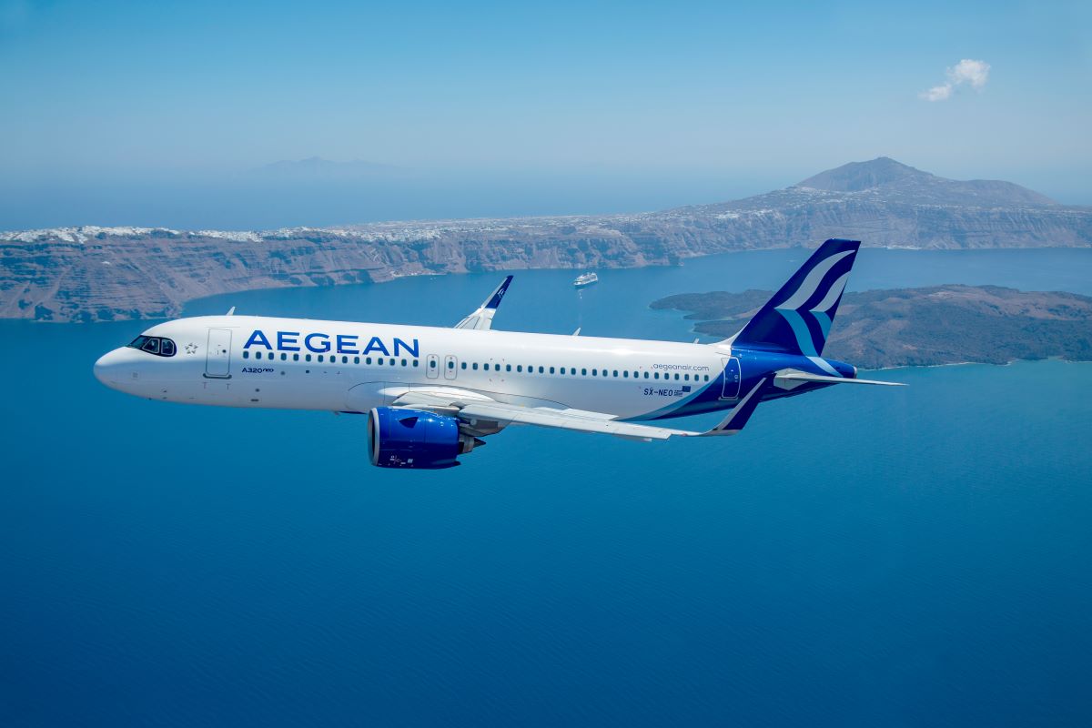 An Aegean Airlines A320neo aircraft mid-flight with a beautiful scenic background containing a large mountain range and an island surrounded by a vast ocean. The sky is bright and clear with few clouds in sight.