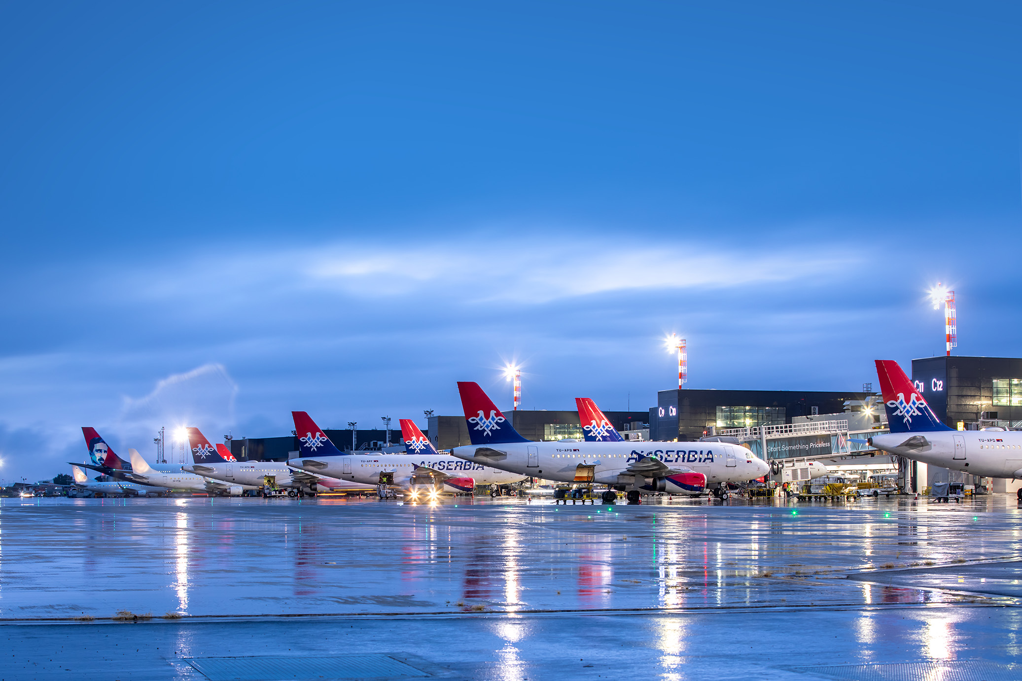 Air Serbia planes are pictured on wet tarmac at airport gates with blue skies and yellow lights.