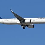 Air Astana Airbus A321neo in flight against a clear blue sky, landing gear down, with a white fuselage and a blue tail bearing a gold emblem.