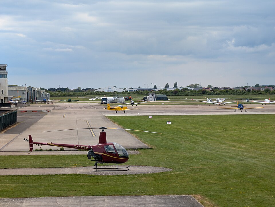 Image shows BCA airport in May 2026 with the control tower to the left, helicopters and small aircraft near the runway on a cloudy day