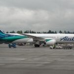 An Alaska Air Boeing 787 Dreamliner plane with blue on white details. The blue is two different shades and runs along the aircraft body to the tail.
