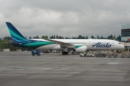 An Alaska Air Boeing 787 Dreamliner plane with blue on white details. The blue is two different shades and runs along the aircraft body to the tail.