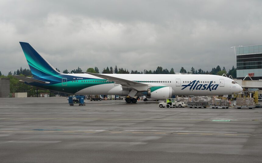 An Alaska Air Boeing 787 Dreamliner plane with blue on white details. The blue is two different shades and runs along the aircraft body to the tail.