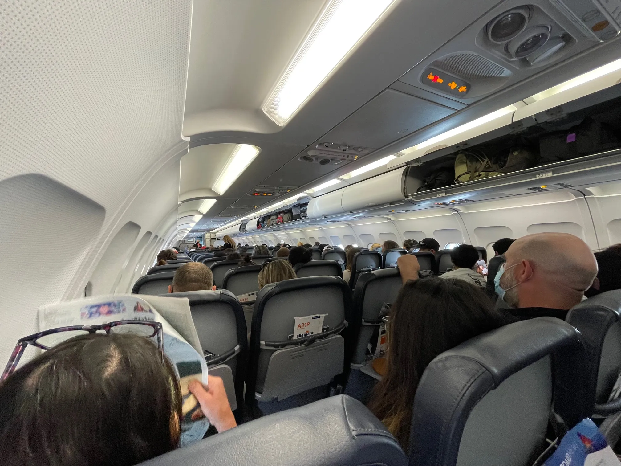 Busy economy cabin seen from a passenger’s seat, with rows of dark grey seats, masked travellers, and open overhead lockers holding bags along the ceiling.