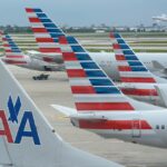 American Airlines aircraft at O'Hare that are parked side-by-side causing the brightly coloured aircraft tails to overlap each other. An aircraft is taking off in the distance into a clear sky.